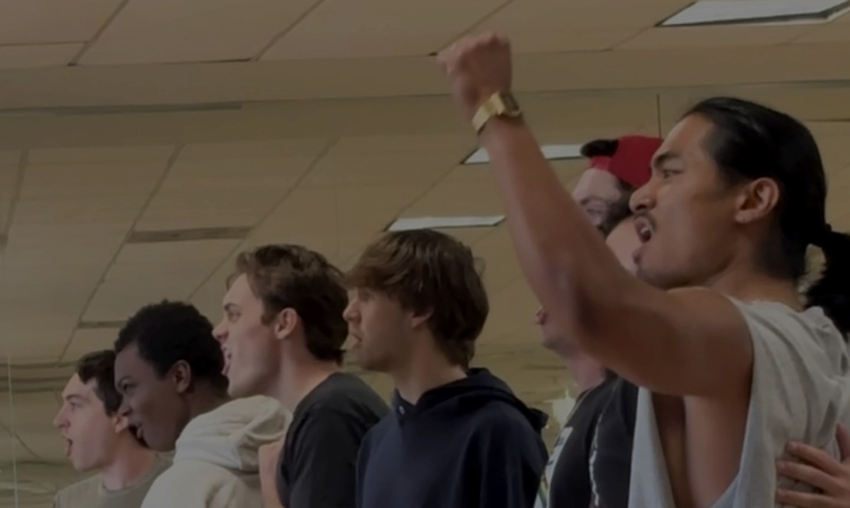 The men of the Les Misérables company singing in the rehearsal room, one with a raised fist